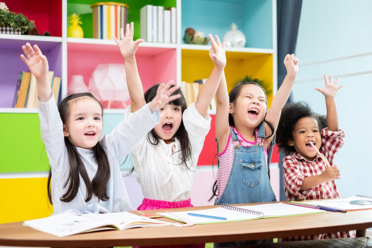 4 Little Girls Raising Their Hands With Enthusiasm Inside A Classroom, Educational Summer Camp For Kids