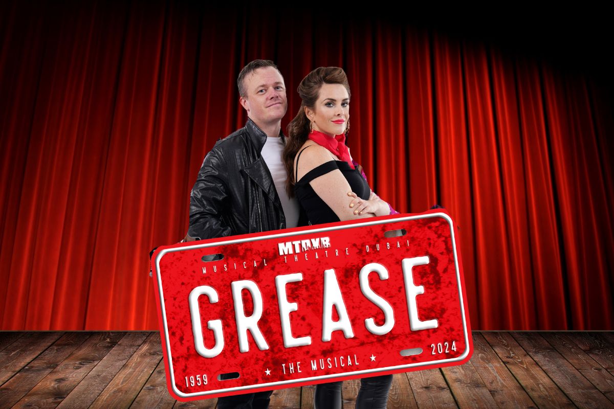 Danny and Sandy posting with a car plate that says GREASE, promotional photo for GREASE the Musical by Musical Theatre Dubai, best things to do in Dubai this Eid Al Adha