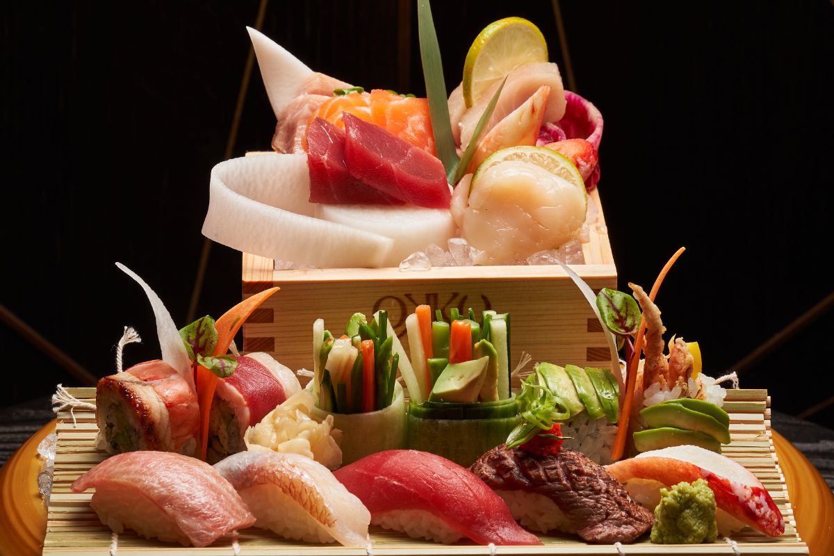 A Variety Of Sushi, Nigiri And Sashimi Well Plated On A Bamboo Crate And Mat. The Variety Includes Tuna, Salmon, Yellowfin, Wagyu Nigiri, Avocado Roll. Octopus Roll, Vegetable Roll, California Roll Well Garnished With Microgreens, Lemon And Vegetables Only At Okku Dubai