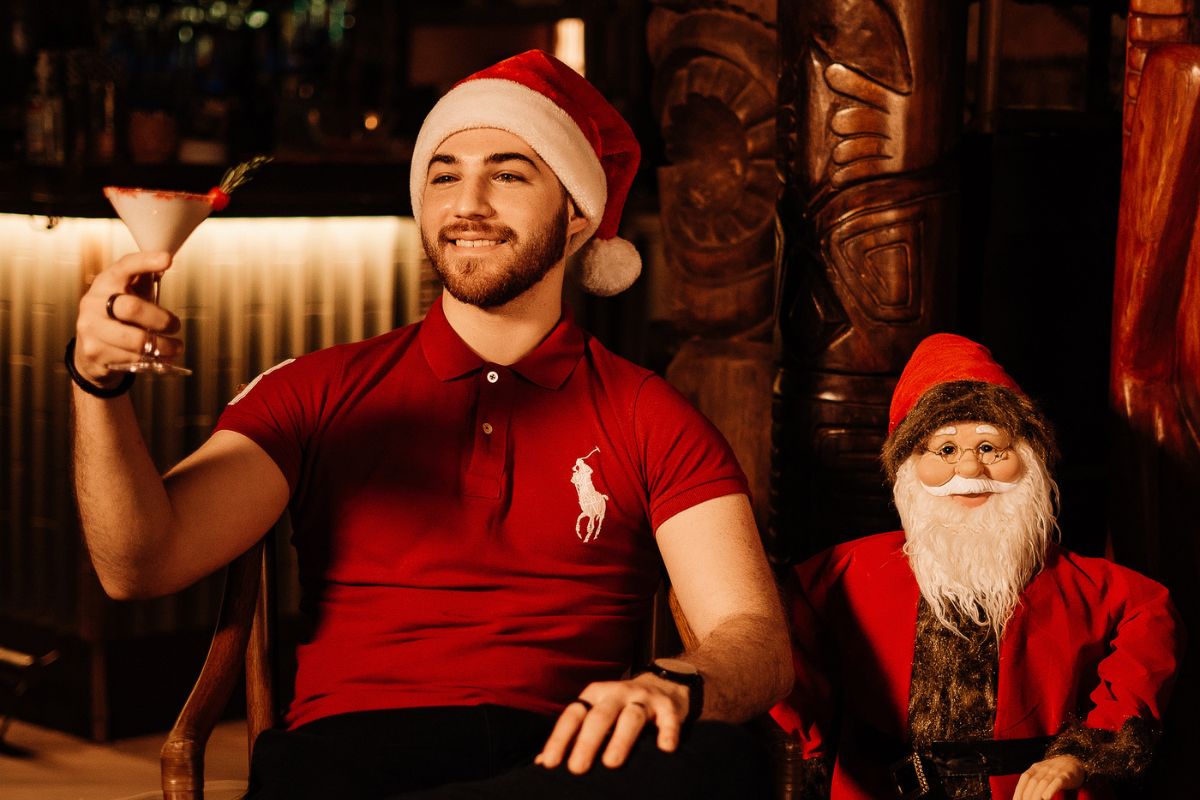 A Guy Holding A Drink With A Santa Hat And Sitting Down Beside A Santa For Christmas In July At Trader Vic'S Dubai