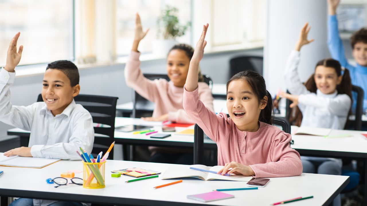Diverse small schoolchildren raising hands at classroom