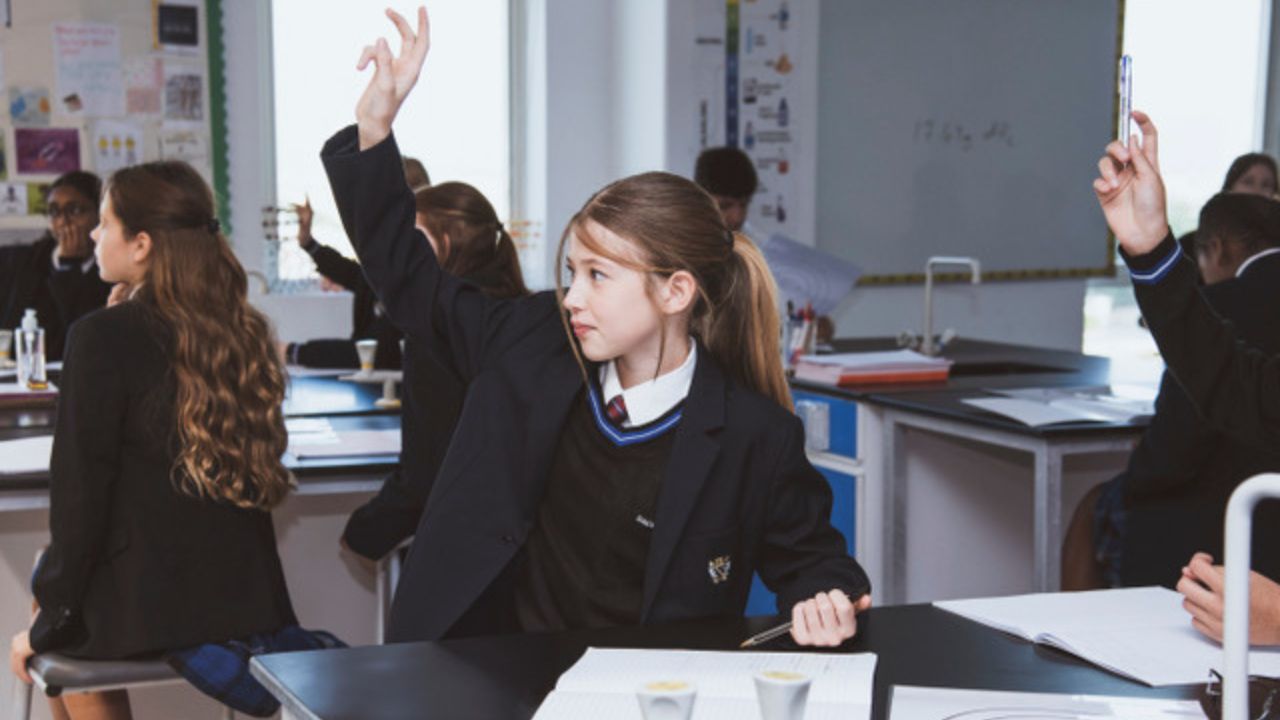 Young girl raising hand in classroom during exam preparation.