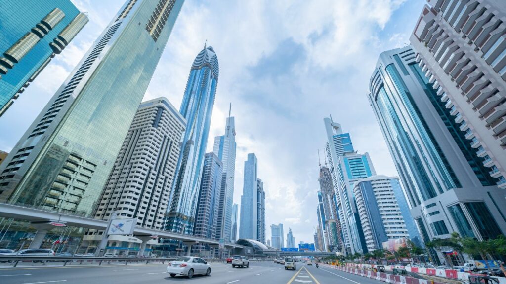 A low angle shot looking up at numerous modern skyscrapers. A wide highway with multiple lanes runs in front of the buildings, with cars driving on it. The cloudy sky suggests daytime. This photo is ideal for urban development projects, travel, and architecture concepts.