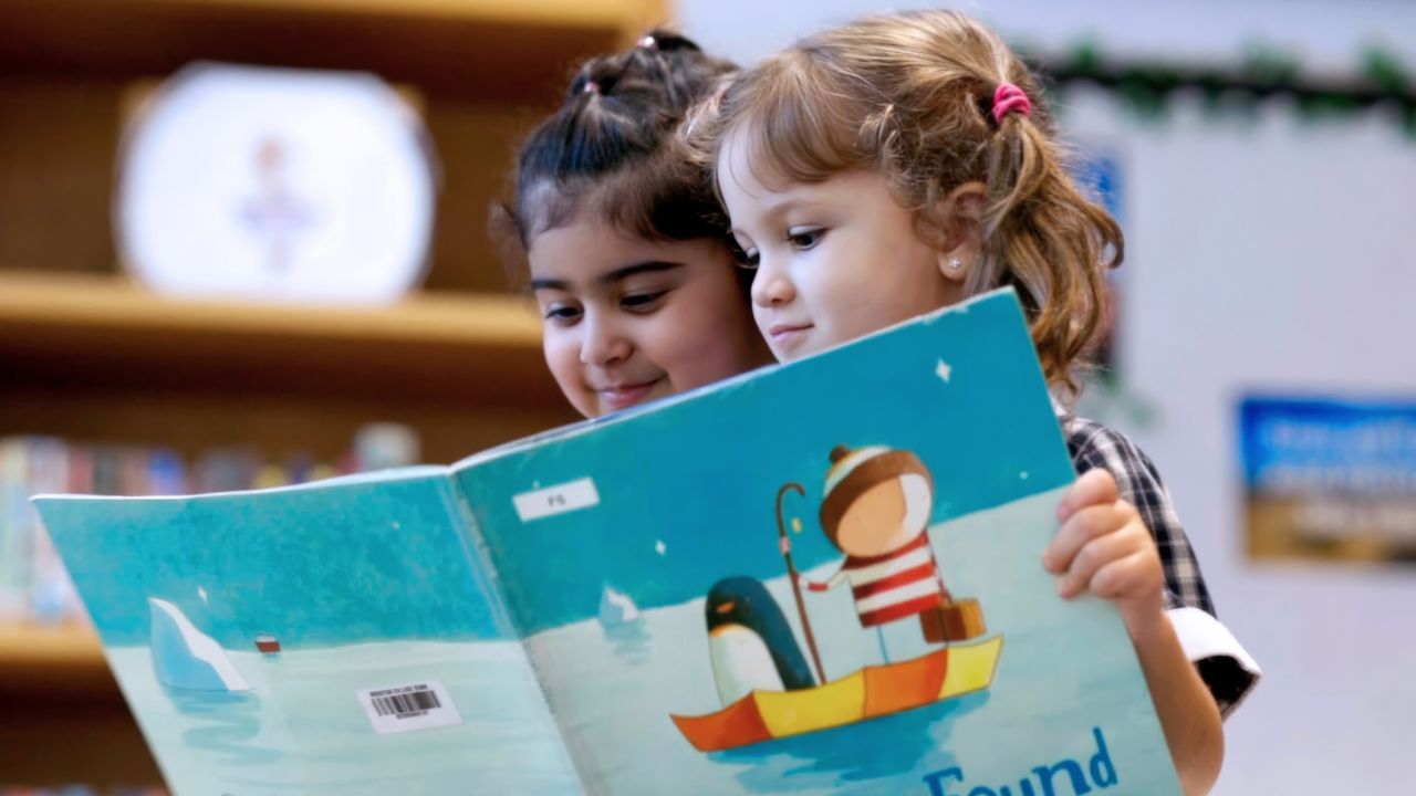 Two young girls reading a colourful children's book in a library setting.