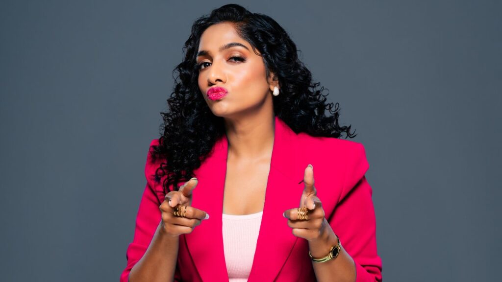 Confident woman with curly hair pointing at camera in pink blazer.