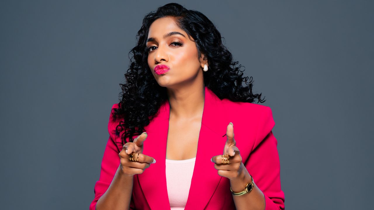 Confident woman with curly hair pointing at camera in pink blazer.