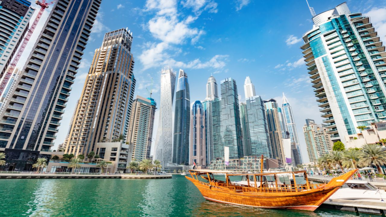 This vibrant cityscape photograph captures a picturesque waterfront scene with sleek modern buildings and a classic wooden boat in view
