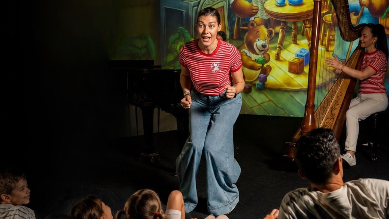 A woman on stage doing a play and another woman at the back playing the harp while audience watch them